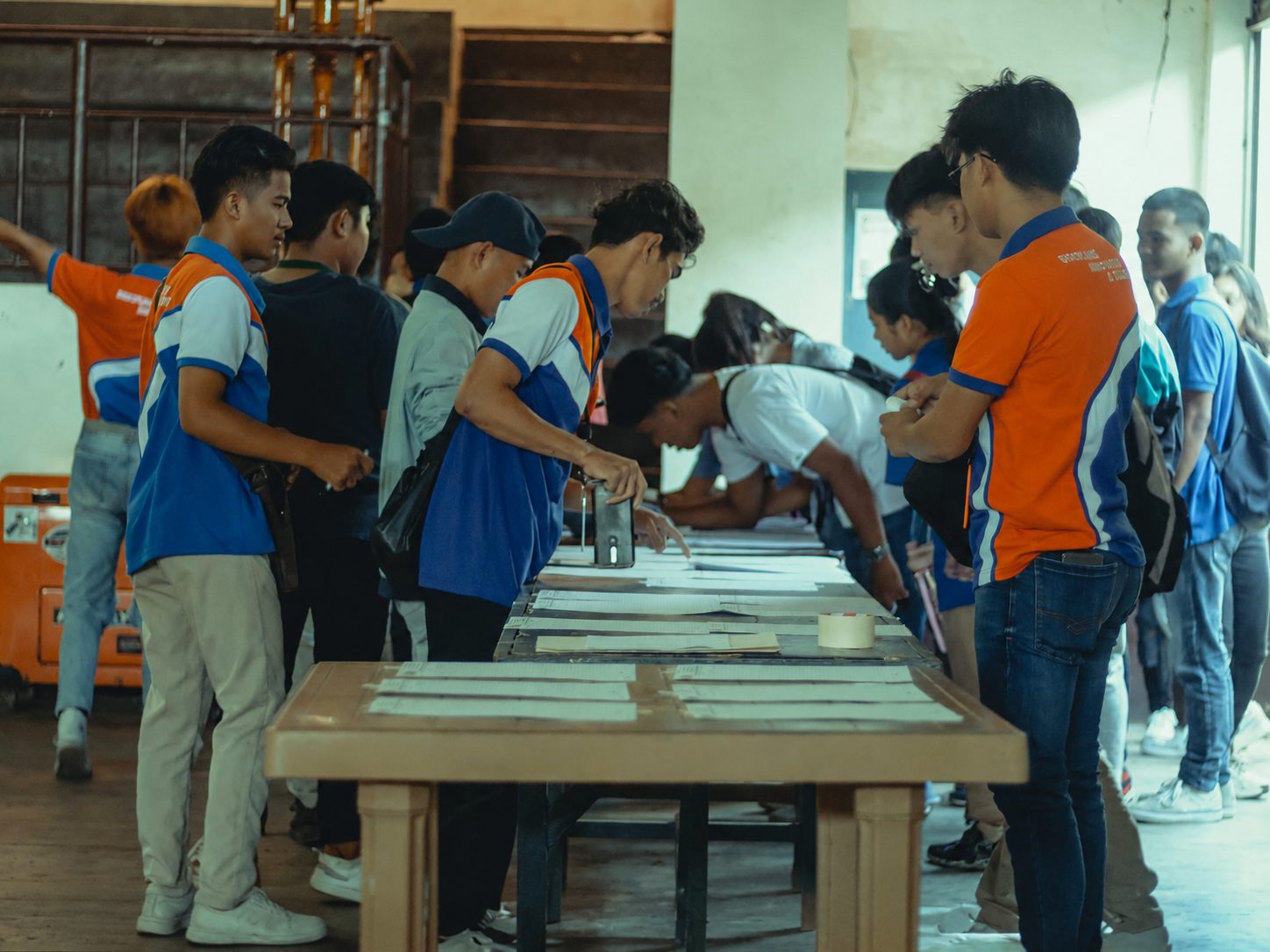 Students queueing at a long table to sign paper attendance sheets